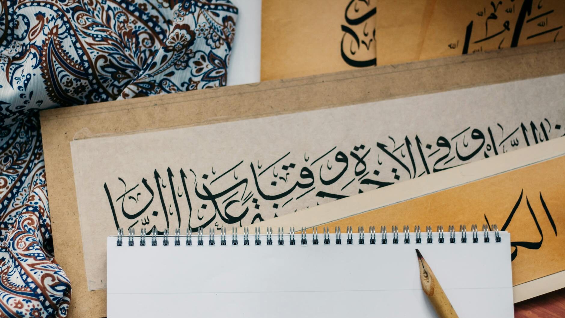 Arabic calligraphy displayed on colorful fabric with a blank notepad and wooden pen.